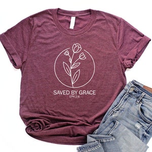 K&ouml;nnte beinhalten: Ein maroonfarbenes T-Shirt mit einer wei&szlig;en Grafik einer Blume in einem Kreis und dem Text "SAVED BY GRACE EPH 2:8".