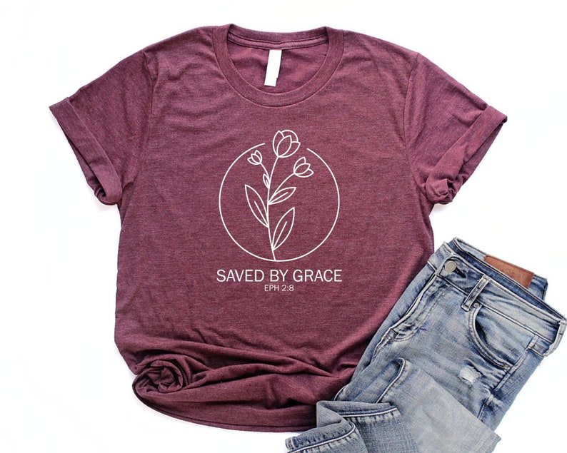 K&ouml;nnte beinhalten: Ein maroonfarbenes T-Shirt mit einer wei&szlig;en Grafik einer Blume in einem Kreis und dem Text "SAVED BY GRACE EPH 2:8".