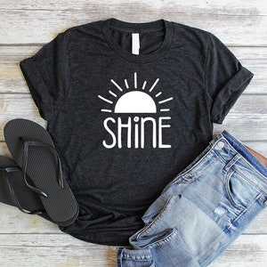 May include: A dark gray t-shirt with a white graphic of a sun with rays and the word "shine" underneath.