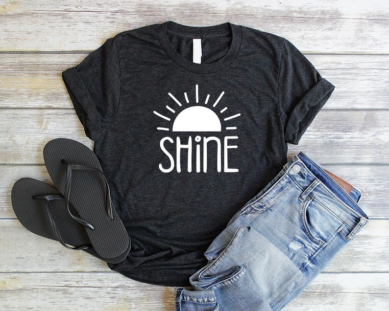 May include: A dark gray t-shirt with a white graphic of a sun with rays and the word "shine" underneath.