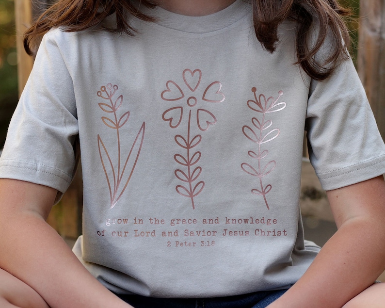 May include: A light gray t-shirt with a rose gold floral design and the text "grow in the grace and knowledge of our Lord and Savior Jesus Christ 2 Peter 3:18".