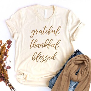 May include: A beige t-shirt with the words "grateful thankful blessed" written in brown cursive lettering.