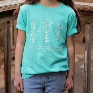 May include: A turquoise blue t-shirt with a white graphic of three stylized flowers and the text "grow in the grace and knowledge of our Lord and Savior Jesus Christ 1 Peter 3:18".