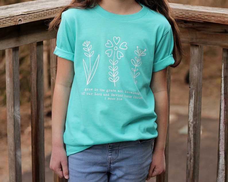 May include: A turquoise blue t-shirt with a white graphic of three stylized flowers and the text "grow in the grace and knowledge of our Lord and Savior Jesus Christ 1 Peter 3:18".