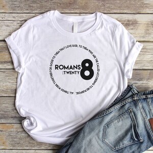 May include: White t-shirt with a black circle graphic featuring the text "Romans : Twenty Eight" and the number "8" in a larger font.