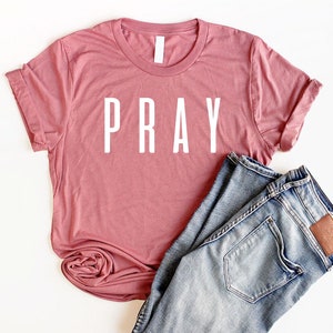May include: A dusty rose colored t-shirt with the word "PRAY" printed in white on the front.