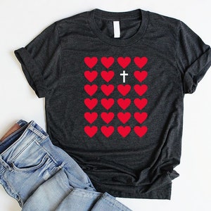 May include: A dark gray t-shirt with a graphic of red hearts and a white cross in the center. The cross is surrounded by hearts.