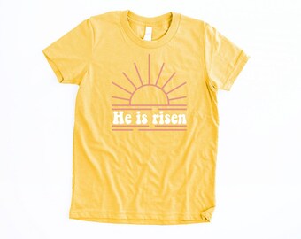 May include: A yellow t-shirt with a pink sun graphic and the text "He is risen" in white.