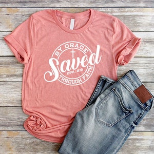 May include: A coral pink t-shirt with a white circle graphic that says "Saved by Grace through Faith" with a cross and the scripture reference "Eph. 2:8".