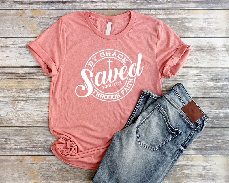 May include: A coral pink t-shirt with a white circle graphic that says "Saved by Grace through Faith" with a cross and the scripture reference "Eph. 2:8".