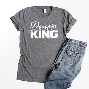 Op de afbeelding: Grijs T-shirt met witte tekst die "Daughter of the King" en "2 Corinthians 6:18" zegt.