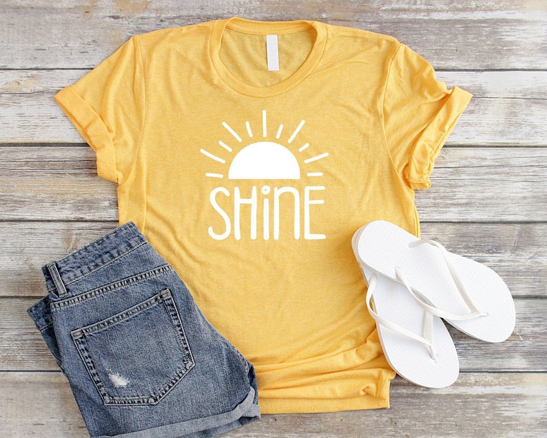 May include: A yellow t-shirt with a white sun and the word "shine" printed on the front. The shirt is paired with denim shorts and white flip flops.