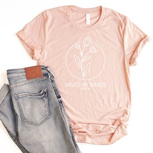 K&ouml;nnte beinhalten: Ein blushfarbenes T-Shirt mit einer wei&szlig;en Umrandung einer Blume und dem Text "SAVED BY GRACE EPH 2:8".