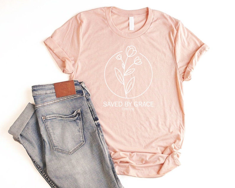 K&ouml;nnte beinhalten: Ein blushfarbenes T-Shirt mit einer wei&szlig;en Umrandung einer Blume und dem Text "SAVED BY GRACE EPH 2:8".