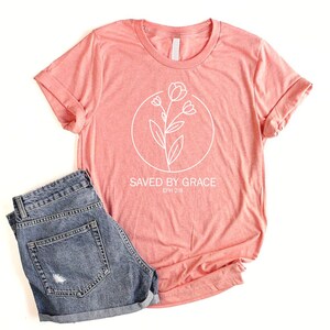 K&ouml;nnte beinhalten: Ein korallenfarbenes T-Shirt mit einer wei&szlig;en Umrandung einer Blume und dem Text "SAVED BY GRACE EPH 2:8".