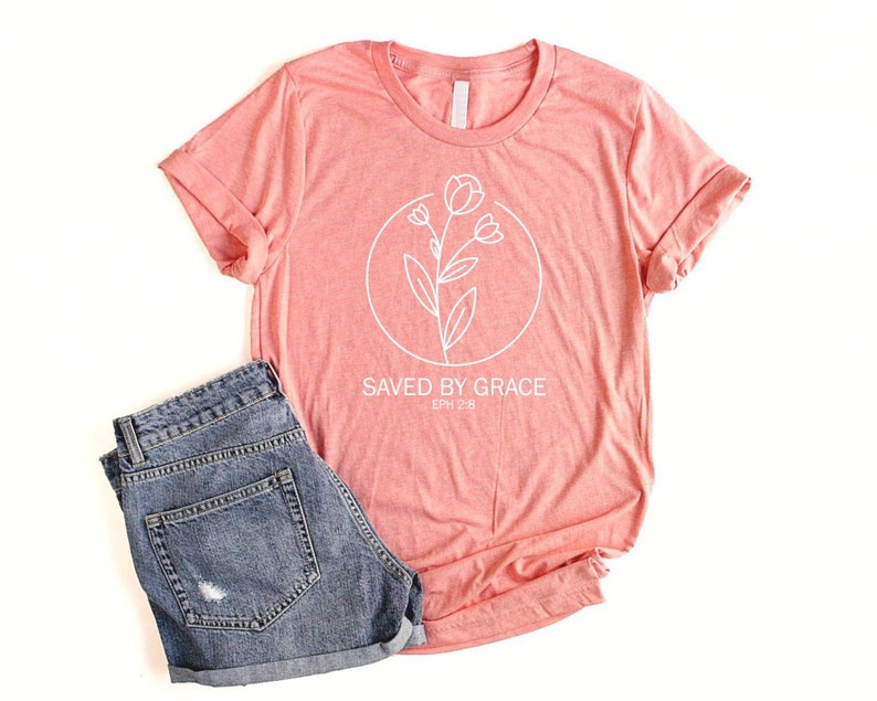 K&ouml;nnte beinhalten: Ein korallenfarbenes T-Shirt mit einer wei&szlig;en Umrandung einer Blume und dem Text "SAVED BY GRACE EPH 2:8".