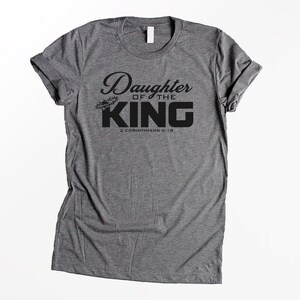 Op de afbeelding: Grijs T-shirt met zwarte tekst die "Daughter of the King" en "2 Corinthians 6:18" zegt.
