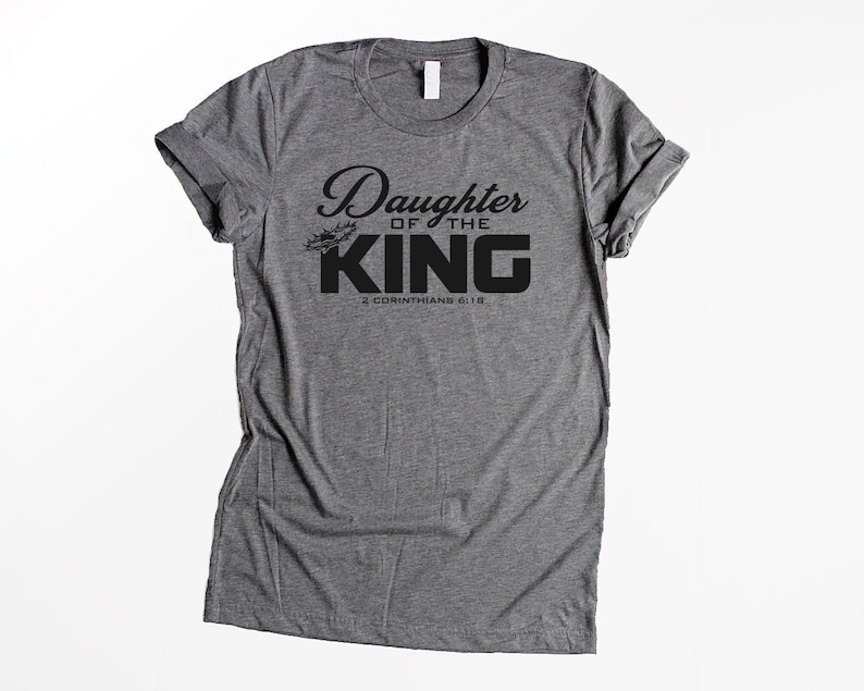 Op de afbeelding: Grijs T-shirt met zwarte tekst die "Daughter of the King" en "2 Corinthians 6:18" zegt.