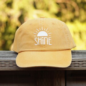 May include: A yellow baseball cap with a white sun and the word "SHINE" embroidered on the front.