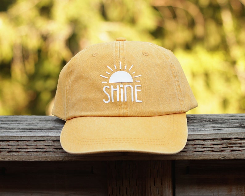 May include: A yellow baseball cap with a white sun and the word "SHINE" embroidered on the front.