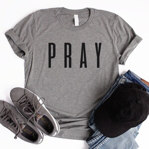 May include: A heather gray t-shirt with the word "PRAY" printed in black on the front. The shirt is laid flat on a white surface with a pair of gray sneakers and a black baseball cap on top of blue jeans.