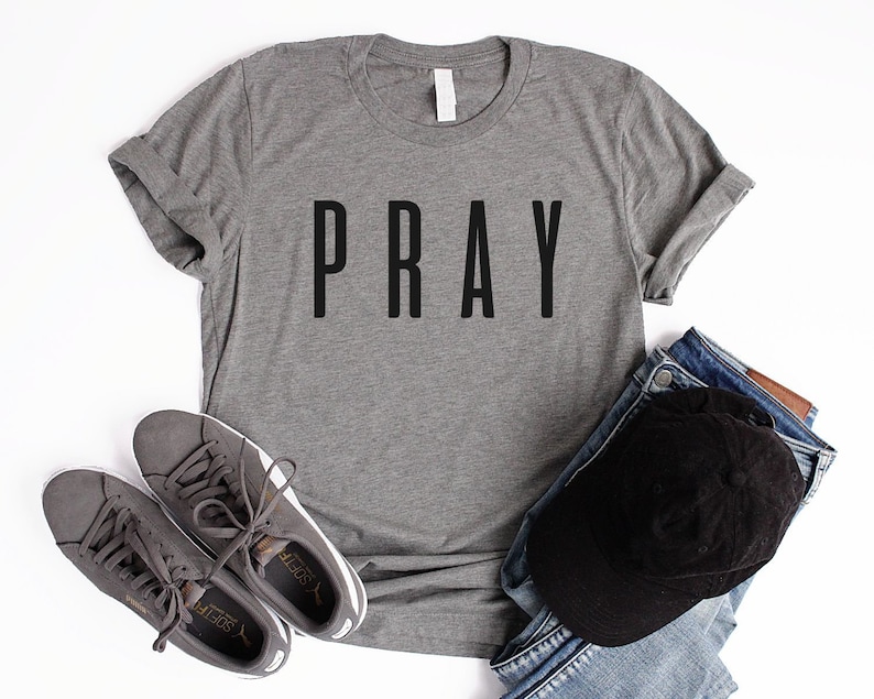 May include: A heather gray t-shirt with the word "PRAY" printed in black on the front. The shirt is laid flat on a white surface with a pair of gray sneakers and a black baseball cap on top of blue jeans.