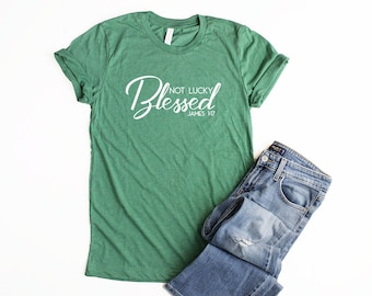 May include: Green t-shirt with white text that reads "Blessed Not Lucky James 1:17". The shirt is folded and laid on a white surface next to a pair of blue jeans.