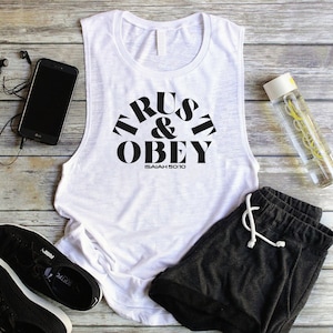 May include: White tank top with black text that reads "TRUST & OBEY ISAIAH 50:10". The tank top is laying on a wooden surface with black athletic shoes, black shorts, and a clear water bottle.