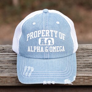 Puede incluir: Una gorra de camionero de mezclilla azul claro con una parte posterior de malla blanca. La gorra tiene un diseño bordado blanco que dice "PROPERTY OF ALPHA & OMEGA" con las letras griegas Alfa y Omega en el centro.