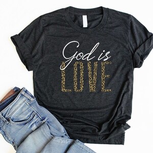 May include: A dark grey t-shirt with the text "God is Love" printed on it. The word "Love" is in a leopard print pattern.