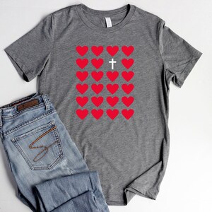 May include: Gray t-shirt with a design of red hearts and a white cross in the center. The hearts are arranged in a grid pattern.