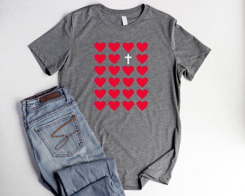 May include: Gray t-shirt with a design of red hearts and a white cross in the center. The hearts are arranged in a grid pattern.