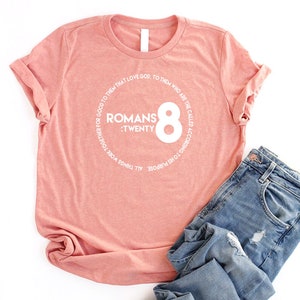 May include: A pink t-shirt with a white circle graphic that says "Romans 8:Twenty" and "All things work together for good to them that love God. To them who are the called according to His purpose."