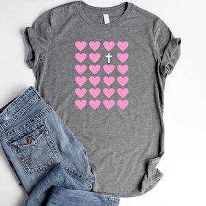May include: A gray t-shirt with a pattern of pink hearts and a white cross in the center of the hearts.