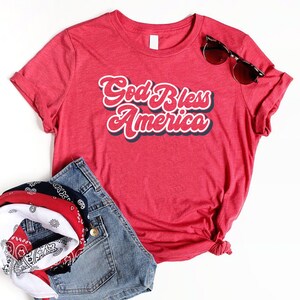 Puede incluir: Una camiseta roja con el texto "God Bless America" en una fuente retro, combinada con pantalones cortos de mezclilla, una bandana y gafas de sol. La camiseta es de cuello redondo.