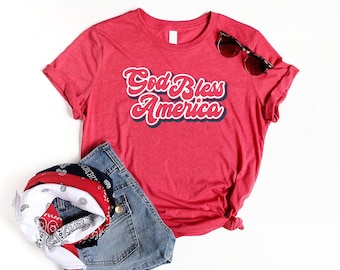 May include: A red t-shirt with the text "God Bless America" in a retro font, paired with denim shorts, a bandana, and sunglasses. The shirt is a crew neck style.
