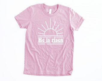 May include: A pink t-shirt with a white graphic of a sunrise and the text "He is risen".
