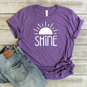 May include: Purple t-shirt with a white sun and the word "SHINE" printed on the front. The shirt is paired with blue jeans.