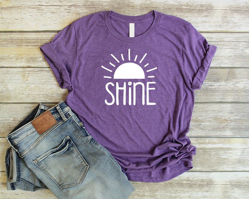 May include: Purple t-shirt with a white sun and the word "SHINE" printed on the front. The shirt is paired with blue jeans.