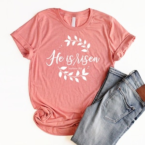 Puede incluir: Una camiseta rosa coral con un gr&aacute;fico blanco que dice "He is risen" con una corona de hojas alrededor del texto y las palabras "Matthew 28:6" debajo. La camiseta se combina con unos vaqueros azules.