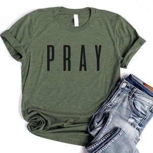 May include: A green t-shirt with the word "PRAY" printed in black on the front. The shirt is rolled up at the sleeves and is laying on a white surface with a pair of blue jeans.