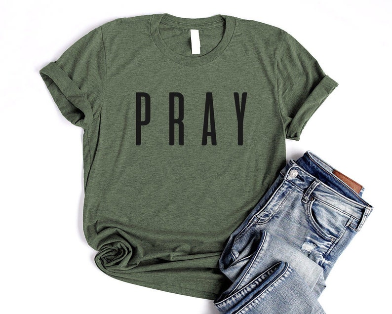 May include: A green t-shirt with the word "PRAY" printed in black on the front. The shirt is rolled up at the sleeves and is laying on a white surface with a pair of blue jeans.
