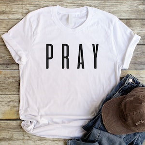 May include: White t-shirt with the word "PRAY" printed in black on the front. The shirt is laying on a wooden surface with a brown baseball cap and blue jeans.