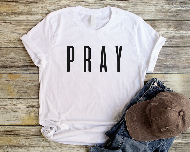 May include: White t-shirt with the word "PRAY" printed in black on the front. The shirt is laying on a wooden surface with a brown baseball cap and blue jeans.