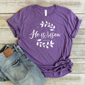 Puede incluir: Camiseta morada con texto blanco que dice "He is risen" y "Matthew 28:6" rodeado de una corona de hojas blancas.