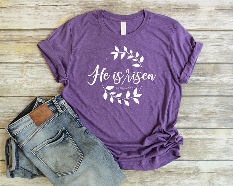 Puede incluir: Camiseta morada con texto blanco que dice "He is risen" y "Matthew 28:6" rodeado de una corona de hojas blancas.