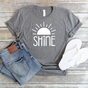 May include: Gray t-shirt with a white sun and the word "shine" printed on it. The shirt is paired with blue jeans and white flip-flops.