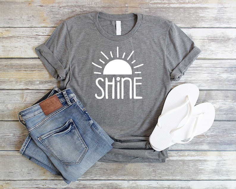 May include: Gray t-shirt with a white sun and the word "shine" printed on it. The shirt is paired with blue jeans and white flip-flops.