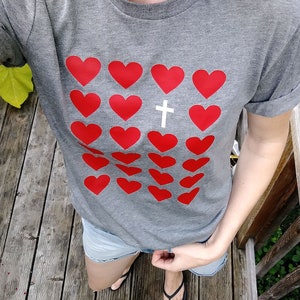 May include: A gray t-shirt with a pattern of red hearts and a white cross in the center. The shirt has a rolled-up sleeve.