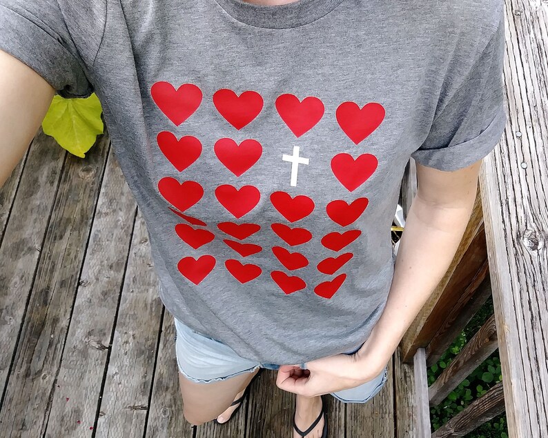 May include: A gray t-shirt with a pattern of red hearts and a white cross in the center. The shirt has a rolled-up sleeve.